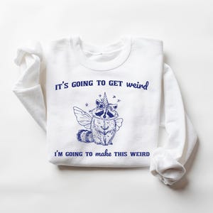 May include: White sweatshirt with blue text that reads "It's going to get weird I'm going to make this weird". A blue illustration of a raccoon with a unicorn horn and wings is in the center of the text.