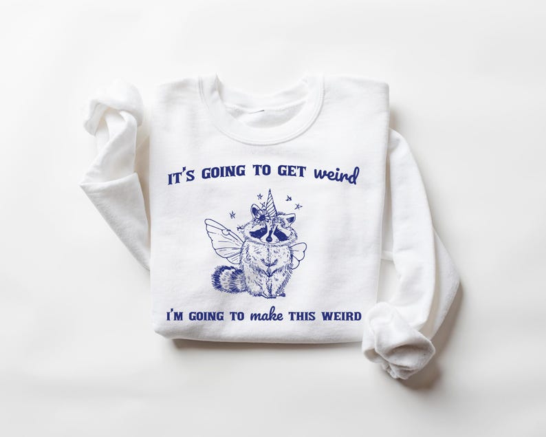 May include: White sweatshirt with blue text that reads "It's going to get weird I'm going to make this weird". A blue illustration of a raccoon with a unicorn horn and wings is in the center of the text.