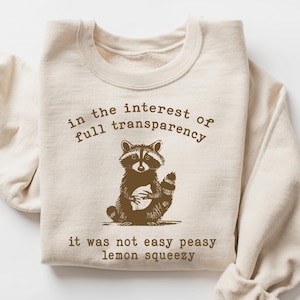 May include: A beige sweatshirt with a brown raccoon graphic and the text "in the interest of full transparency it was not easy peasy lemon squeezy".