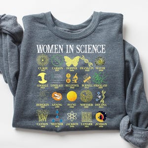 Pode incluir: Uma camisa de moletom cinza escura com o texto "WOMEN IN SCIENCE" impresso em branco. A camisa de moletom apresenta um design gráfico com ilustrações de mulheres na ciência e seus nomes e anos de nascimento.