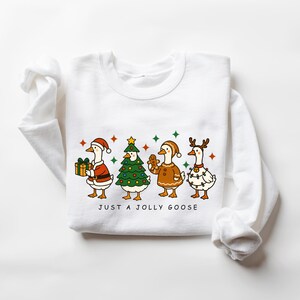 May include: White sweatshirt featuring a festive illustration of four geese dressed for Christmas. One goose holds a gift, another is a Christmas tree, a third is a gingerbread cookie, and the last is decorated with lights. The text "JUST A JOLLY GOOSE" is below the geese.