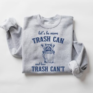Pu&ograve; includere: Felpa grigia con una grafica blu di un procione in un bidone della spazzatura. Il testo recita "let's be more trash can and less trash can't".