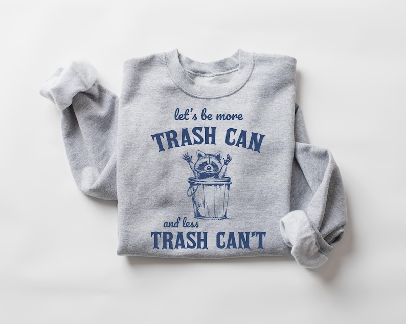 Pu&ograve; includere: Felpa grigia con una grafica blu di un procione in un bidone della spazzatura. Il testo recita "let's be more trash can and less trash can't".