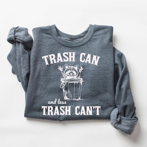 Pu&ograve; includere: Una felpa grigia scura con una grafica bianca di un procione in un bidone della spazzatura. Il testo recita "Trash Can and less Trash Can't".