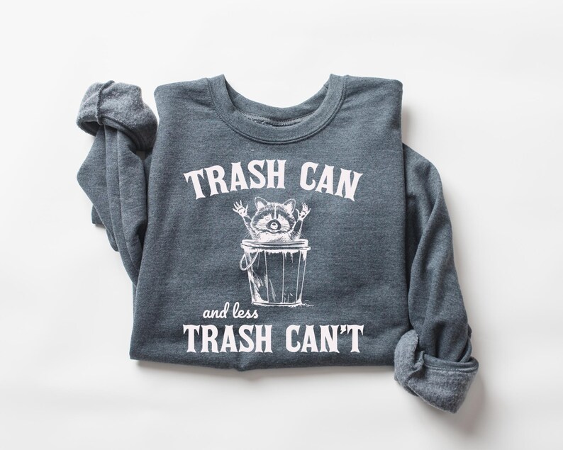 Pu&ograve; includere: Una felpa grigia scura con una grafica bianca di un procione in un bidone della spazzatura. Il testo recita "Trash Can and less Trash Can't".
