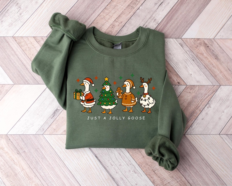 May include: Green crewneck sweatshirt featuring a festive design with four cartoon geese dressed in Christmas attire. The geese are wearing Santa hats, a Christmas tree costume, a gingerbread outfit, and reindeer antlers. The text "JUST A JOLLY GOOSE" is printed below the geese.