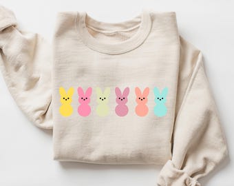 Peut inclure: Un sweat-shirt beige avec une rangée de six silhouettes de lapins de couleur pastel. Les lapins sont jaunes, roses, vert clair, bleu clair, violets et orange.