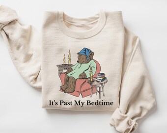 Può includere: Una felpa beige con una grafica di un orso che indossa un berretto da notte blu e siede su una poltrona rossa. L'orso tiene un libro e ci sono delle candele su un tavolo accanto alla poltrona. Il testo "It's Past My Bedtime" è stampato sotto la grafica.