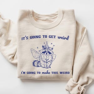 May include: A beige sweatshirt with the text "It's going to get weird" and "I'm going to make this weird" in blue. A cartoon raccoon with wings and a unicorn horn is in the center of the sweatshirt.