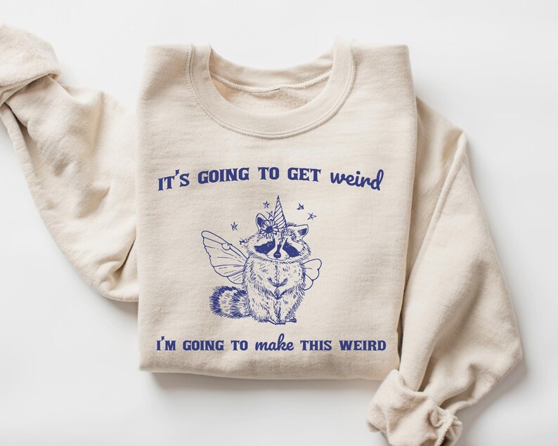 May include: A beige sweatshirt with the text "It's going to get weird" and "I'm going to make this weird" in blue. A cartoon raccoon with wings and a unicorn horn is in the center of the sweatshirt.