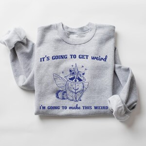 May include: Gray sweatshirt with a blue graphic of a raccoon wearing a unicorn horn and wings. The text "It's going to get weird" and "I'm going to make this weird" is printed on the sweatshirt.