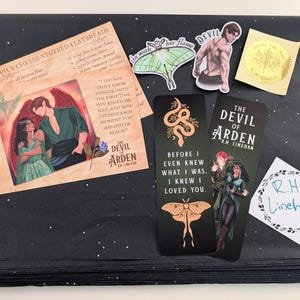 May include: A collection of items on a black surface with white star-like accents. Includes a recipe card, stickers, and bookmarks featuring illustrations and text. The bookmarks and recipe card have the text "The Devil of Arden".