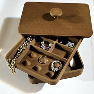 Może przedstawiać: Brązowe pudełko na biżuterię z otwieraną pokrywą i gałką. Otwarta skrzynka odsłania wiele przegródek zawierających biżuterię, w tym pierścionki, kolczyki i złoty łańcuszek. Pudełko wykonane jest z teksturowanego materiału.