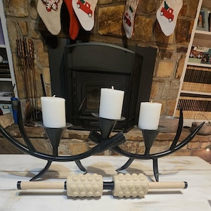 May include: A black metal candle holder with three white candles, and two massage rollers with wooden handles and textured beige rollers. Christmas stockings hang above a fireplace.