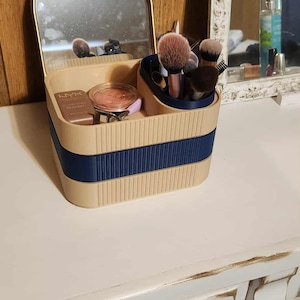 May include: A beige and navy blue makeup organizer with a built-in mirror. The organizer holds makeup brushes, blush, and other cosmetics. The organizer sits on a white dresser with dark knobs.