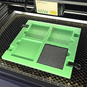 May include: A green laser cutting mat with four square cutouts and a black slate tile in one of the cutouts. The mat is on a honeycomb-patterned surface. The laser cutter is visible above the mat, with the text "XTOOL 40W" on the front.