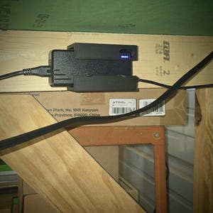 May include: A black power adapter with a blue light indicator is mounted to a wooden surface. The adapter is plugged into a black power cord. The text on the wooden surface reads "XTOOL" and "on iPark, No. 1001 Xueyuan Province, 518000, China".