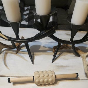 May include: A black metal candelabra with three white candles, antler accents, and a beige massage roller with wooden handles and a textured surface. The candelabra is on a white wooden surface.