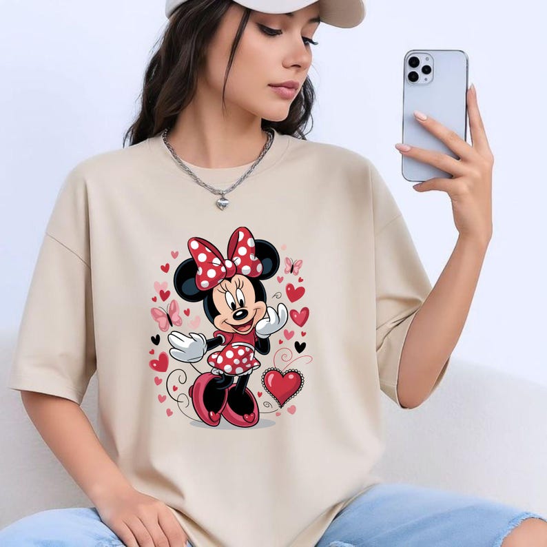 May include: A beige short-sleeved t-shirt featuring a graphic print of Minnie Mouse wearing a red and white polka dot dress and bow. The design is surrounded by various red and pink hearts, black hearts, and small butterflies in a decorative arrangement.