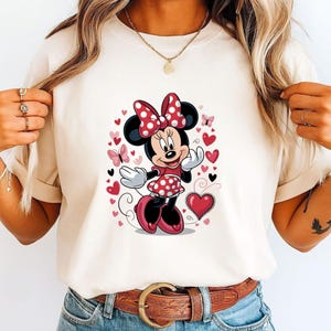 May include: A cream-colored t-shirt featuring a graphic print of a cartoon mouse character wearing a red and white polka dot dress and matching bow. The design is surrounded by various red and pink hearts and small butterflies on the front.