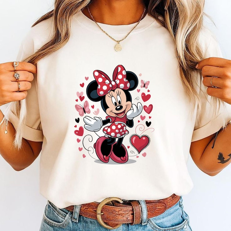 May include: A cream-colored t-shirt featuring a graphic print of a cartoon mouse character wearing a red and white polka dot dress and matching bow. The design is surrounded by various red and pink hearts and small butterflies on the front.