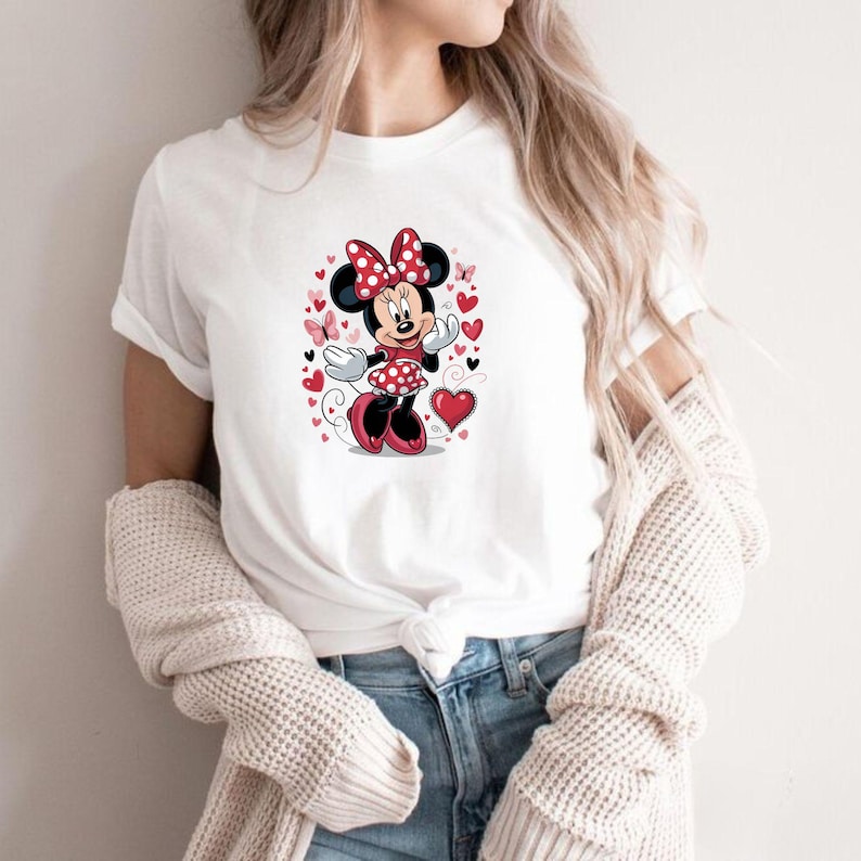 May include: A white short-sleeved t-shirt featuring a graphic print of a cartoon mouse character wearing a red and white polka dot dress and matching bow, surrounded by red and black hearts and small pink butterflies on the front center.
