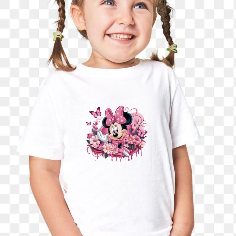 May include: White t-shirt featuring a graphic of Minnie Mouse in a pink dress and bow, surrounded by pink flowers and butterflies. The design has a watercolor effect with dripping paint details.