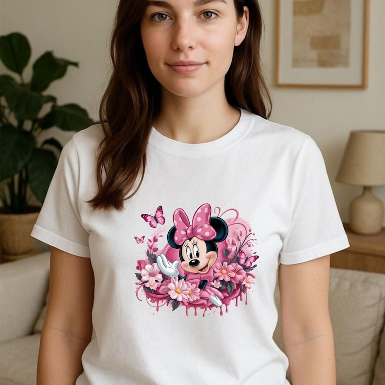 May include: White t-shirt featuring a Minnie Mouse graphic. The design includes Minnie Mouse wearing a pink polka-dot bow and dress, surrounded by pink flowers and butterflies. The graphic is centered on the shirt.