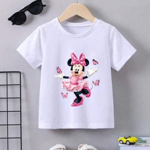 May include: A white short-sleeved t-shirt featuring a graphic print of Minnie Mouse wearing a pink polka-dot dress and bow, surrounded by several pink butterflies. The shirt has a round neckline and is displayed on a light-colored hanger.