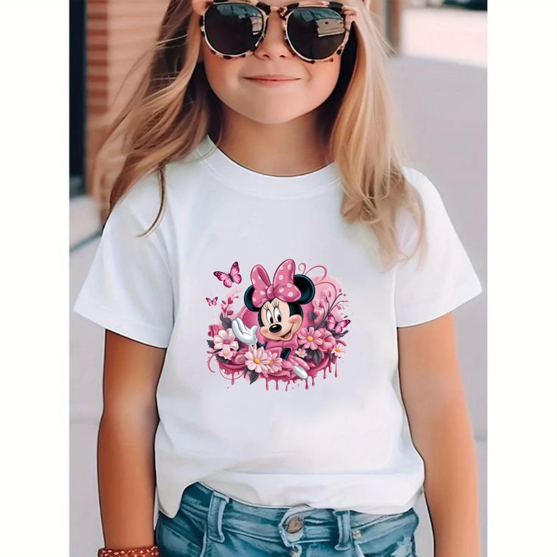 May include: White t-shirt featuring a Minnie Mouse graphic. The design includes Minnie Mouse with a pink bow, surrounded by pink flowers and butterflies. The shirt is worn by a person.