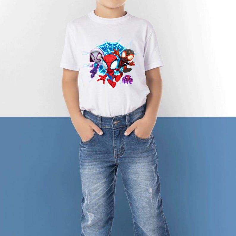 May include: White t-shirt with a vibrant graphic of Spider-Man and two other characters. The design includes a blue spiderweb and action poses. The child is wearing blue denim jeans.