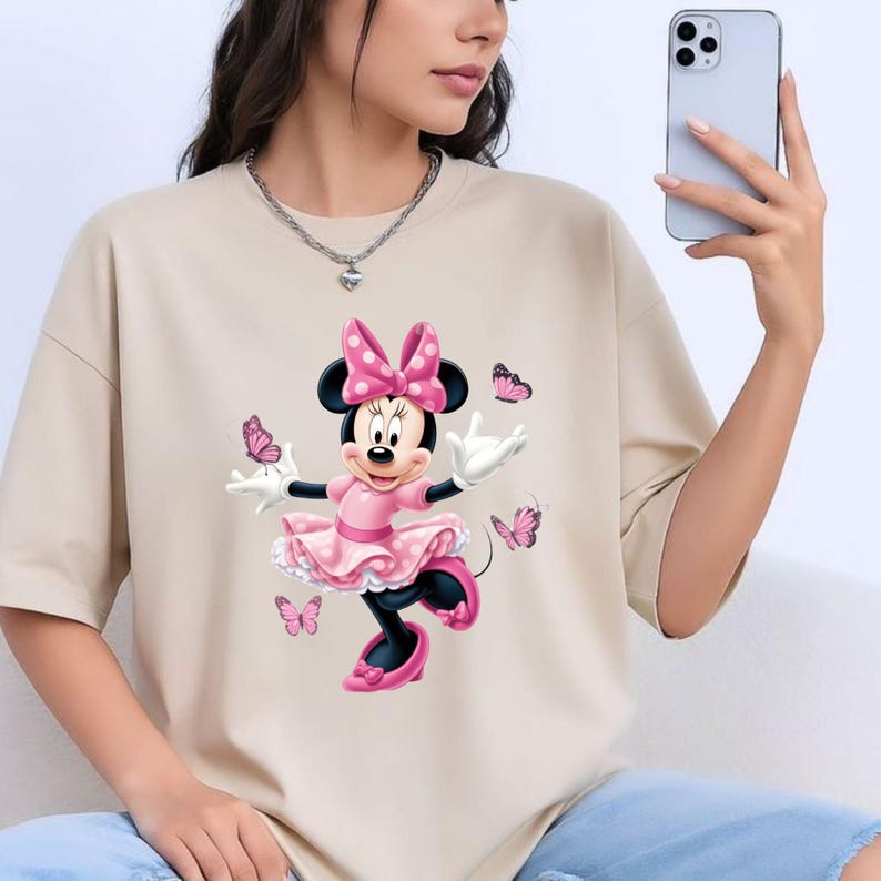 May include: A beige short-sleeved t-shirt featuring a graphic print of a cartoon mouse wearing a pink polka-dot dress and bow, surrounded by several small pink butterflies. The mouse has white gloves and pink shoes, set against a plain background.
