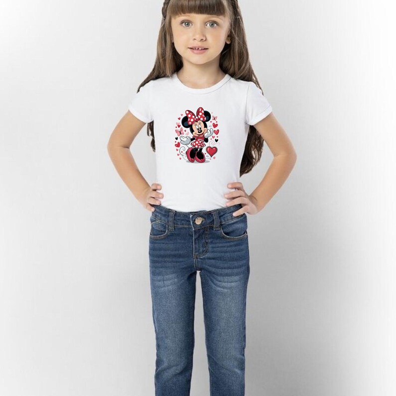 May include: A white short-sleeved t-shirt featuring a graphic print of Minnie Mouse surrounded by red hearts and small butterflies, paired with dark blue denim jeans. The character wears a red polka-dot dress and matching bow.