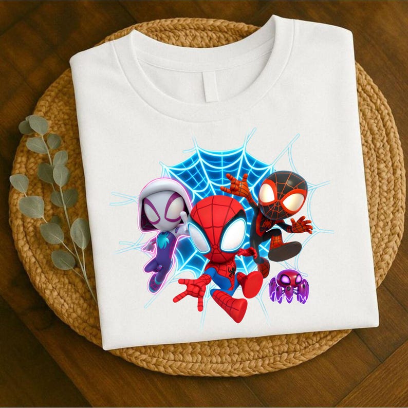 May include: White t-shirt with a graphic of three cartoon Spider-Man characters and a purple spider against a blue web. The t-shirt is folded on a woven mat.