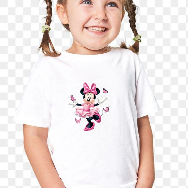 May include: A white short-sleeved t-shirt featuring a graphic print of Minnie Mouse wearing a pink polka-dot dress and matching bow, surrounded by several small pink butterflies. The character is depicted in a standing pose with arms outstretched.