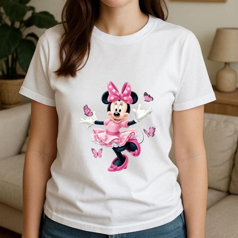 May include: A white short-sleeved t-shirt featuring a graphic print of Minnie Mouse wearing a pink polka-dot dress and bow, surrounded by several small pink butterflies. The character is depicted with white gloves and pink shoes on a plain background.