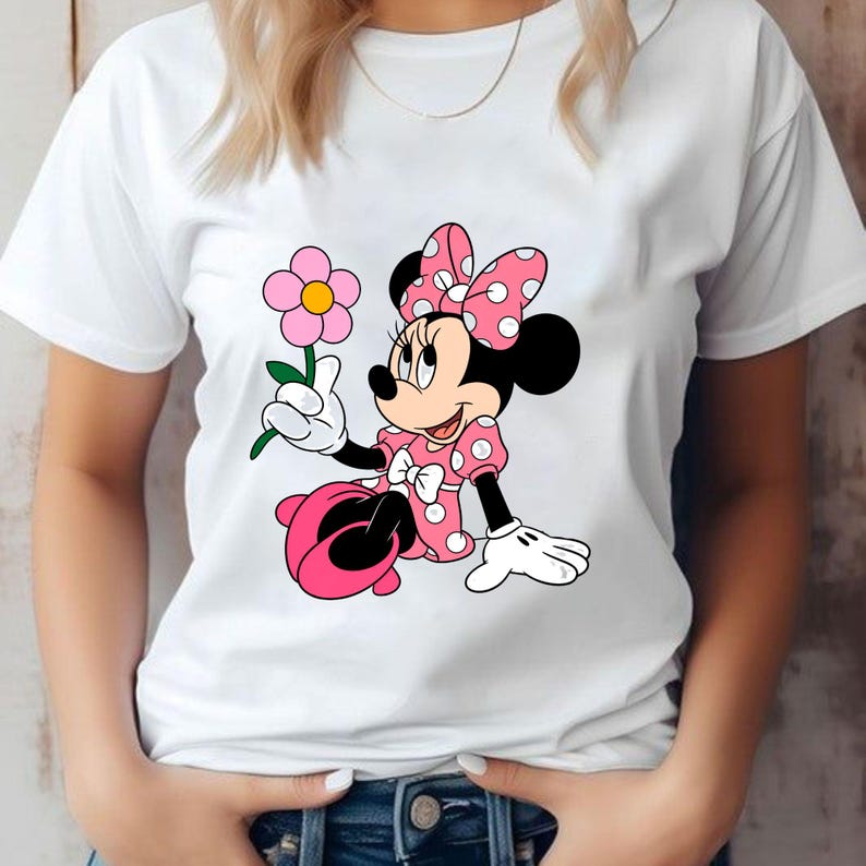 May include: A white short-sleeved t-shirt featuring a graphic print of Minnie Mouse wearing a pink polka-dot dress and a matching bow. The character is depicted sitting down and holding a single pink flower with a yellow center in one hand.