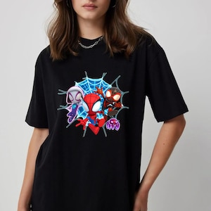 May include: Black t-shirt with a vibrant graphic of three Spider-Man characters set within a blue web. The characters are in red, black, and white costumes. A purple spider is also included in the design. The shirt is made of a soft material.