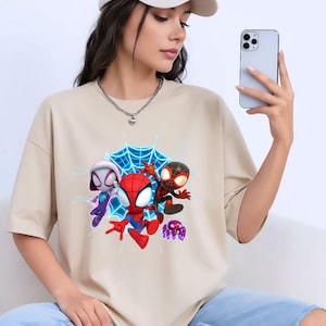 May include: Beige t-shirt with a vibrant graphic of three Spider-Man characters and a purple spider against a blue web. The design includes Spider-Gwen, Spider-Man, and Miles Morales.