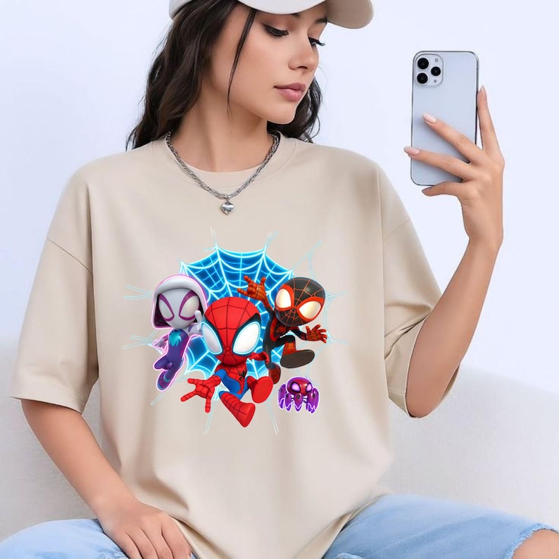 May include: Beige t-shirt with a vibrant graphic of three Spider-Man characters and a purple spider against a blue web. The design includes Spider-Gwen, Spider-Man, and Miles Morales.