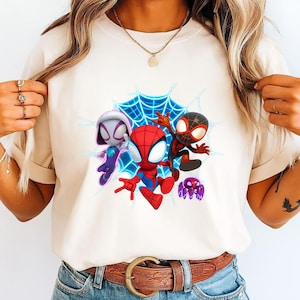 May include: A cream-coloured t-shirt with a colourful graphic of three Spider-Man characters and a purple spider against a blue web background. The Spider-Man characters are in red, black, and white, with blue and purple accents.