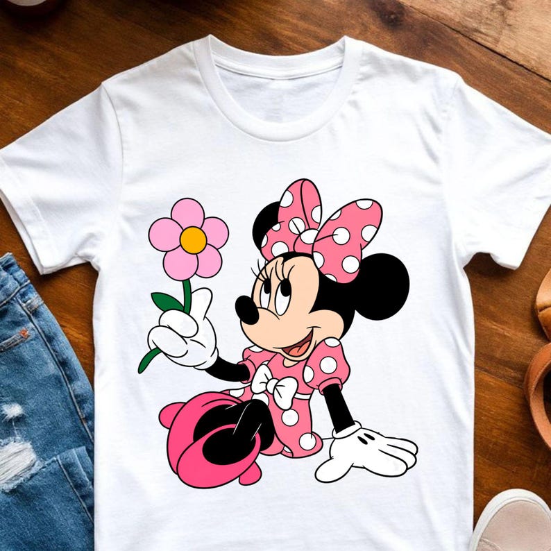 May include: A white short-sleeved t-shirt featuring a graphic print of Minnie Mouse wearing a pink polka-dot dress and matching bow. The character is depicted sitting down and holding a single pink flower with a yellow center and green stem.