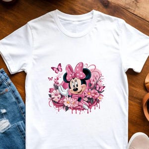 May include: White t-shirt featuring a colorful graphic of Minnie Mouse in a pink dress and bow, surrounded by pink flowers and butterflies. The design has a watercolor effect, with dripping paint details.