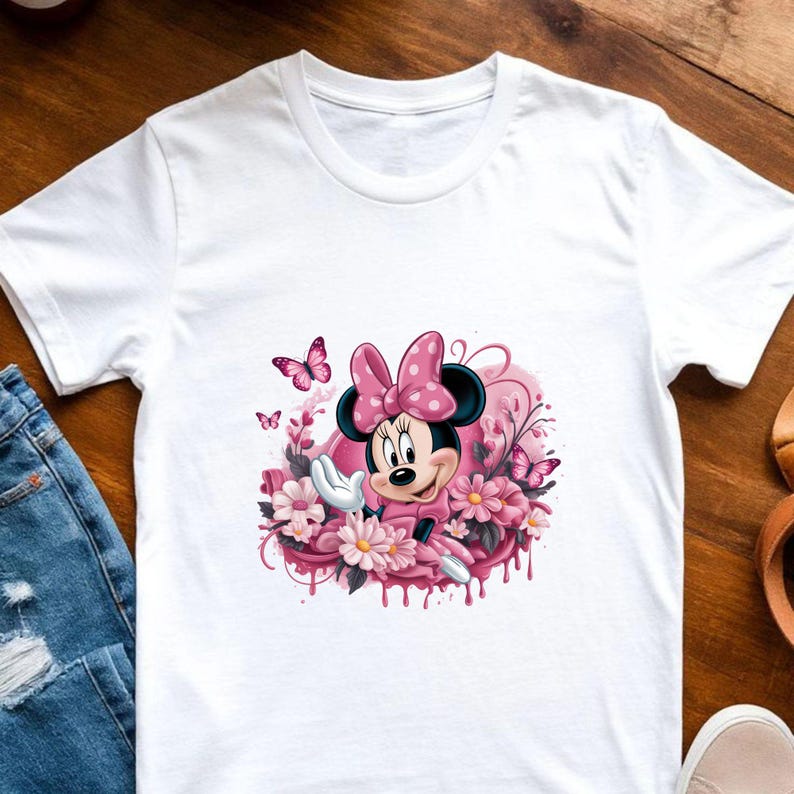 May include: White t-shirt featuring a colorful graphic of Minnie Mouse in a pink dress and bow, surrounded by pink flowers and butterflies. The design has a watercolor effect, with dripping paint details.