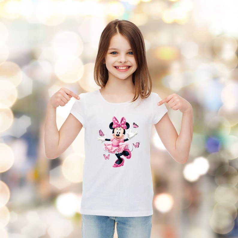 May include: A white short-sleeved t-shirt featuring a graphic print of Minnie Mouse wearing a pink dress and a matching bow, surrounded by several small pink butterflies. The character is depicted in a dynamic pose with arms outstretched.