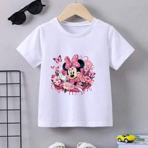 May include: White t-shirt featuring a colorful graphic of Minnie Mouse surrounded by pink flowers and butterflies. Minnie Mouse is wearing a pink dress and a polka-dot bow. The shirt is hanging on a wooden hanger.