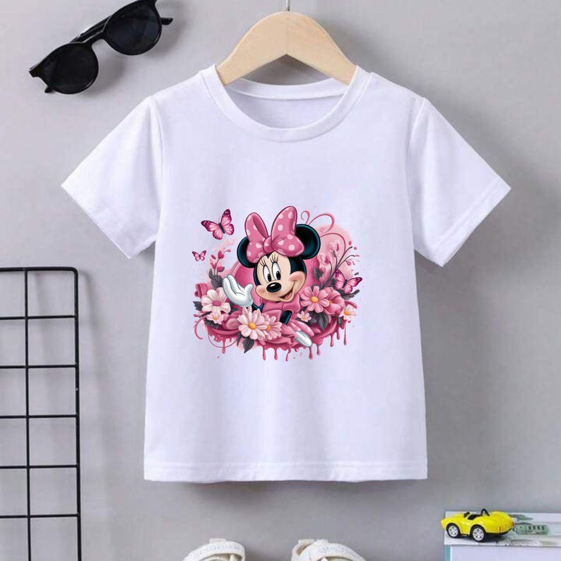 May include: White t-shirt featuring a colorful graphic of Minnie Mouse surrounded by pink flowers and butterflies. Minnie Mouse is wearing a pink dress and a polka-dot bow. The shirt is hanging on a wooden hanger.