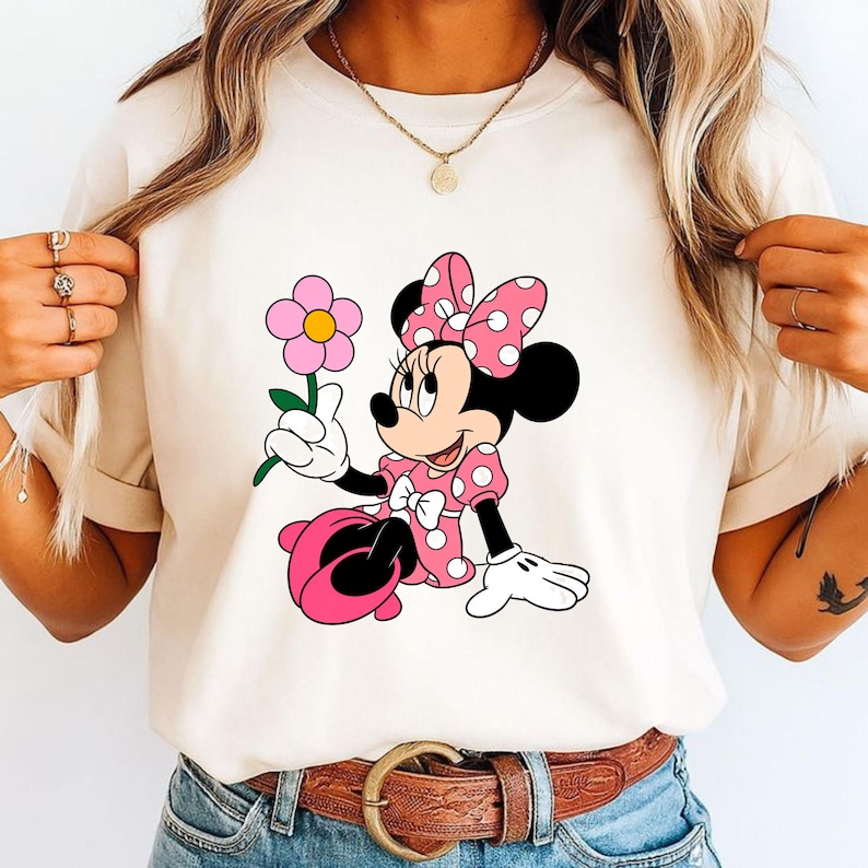 May include: A cream-colored short-sleeve t-shirt featuring a graphic print of Minnie Mouse wearing a pink polka-dot dress and matching bow, holding a pink flower with a yellow center. The character has black ears, white gloves, and pink shoes.