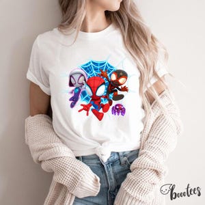 May include: White t-shirt with a vibrant graphic of three Spider-Man characters and a purple spider against a blue web. The tee has a crew neck and short sleeves. The brand name "bootees" is printed on the bottom right.