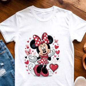 May include: A white short-sleeved t-shirt featuring a graphic print of Minnie Mouse wearing a red and white polka-dot dress and matching bow. The design is surrounded by various red and pink hearts and small butterflies on the front center.